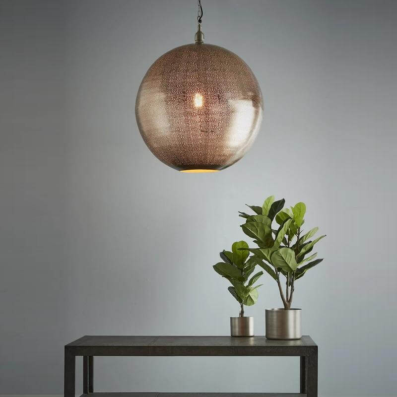 A modern pendant light with a nickel finish and small perforations in the shade, suspended over a grey table, featuring two plants and a geometric metal leg.