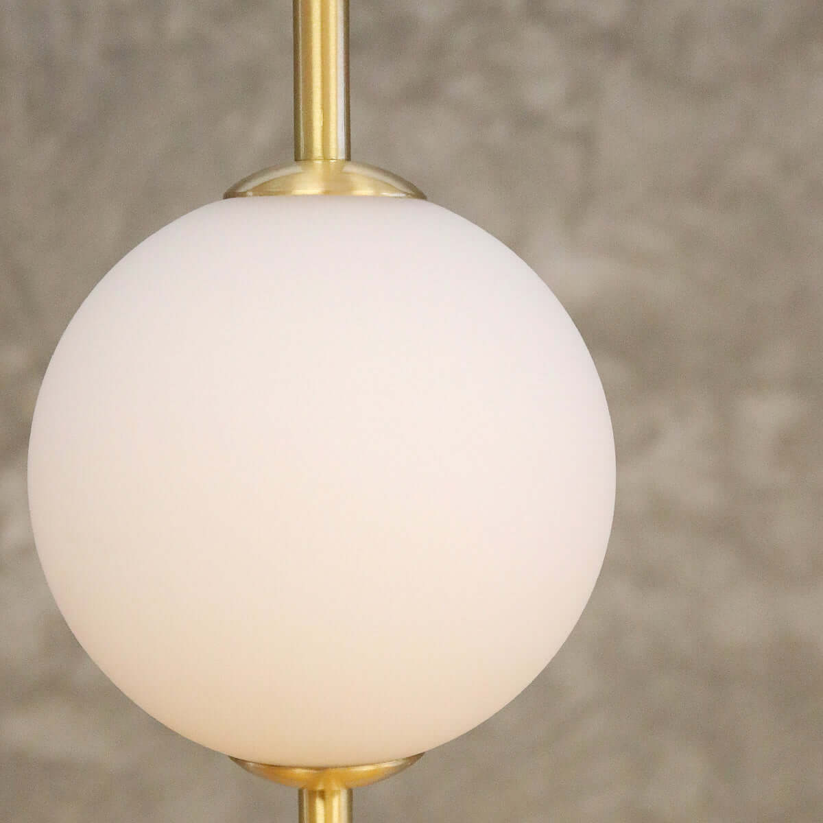 A modern pendant light with a glowing white spherical shade and brushed brass vertical rods, suspended against a textured concrete wall.