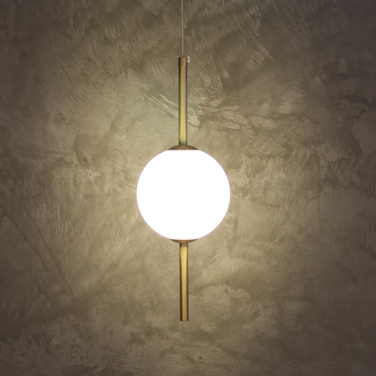A modern pendant light with a glowing white spherical shade and brushed brass vertical rods, suspended against a textured concrete wall.
