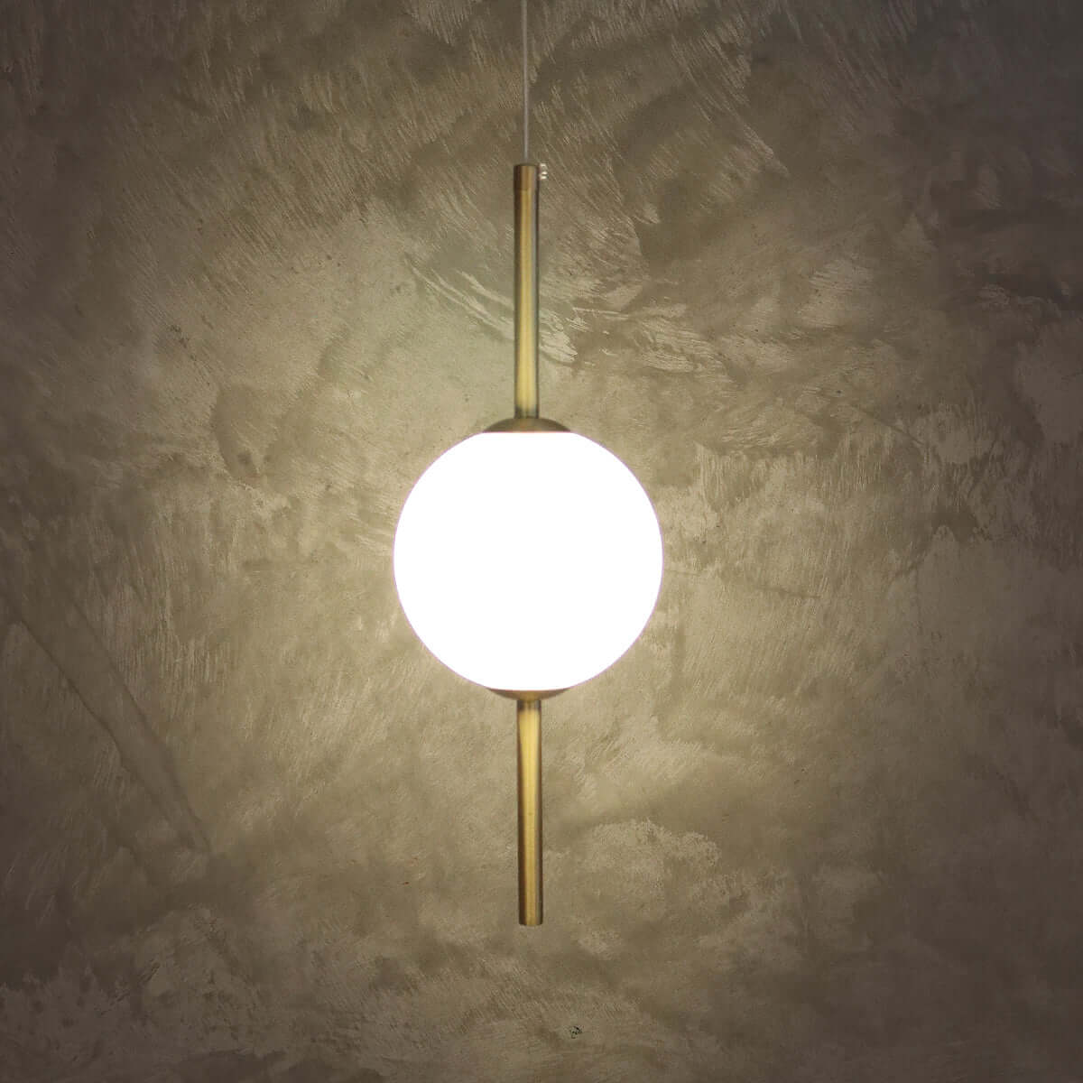 A modern pendant light with a glowing white spherical shade and brushed brass vertical rods, suspended against a textured concrete wall.