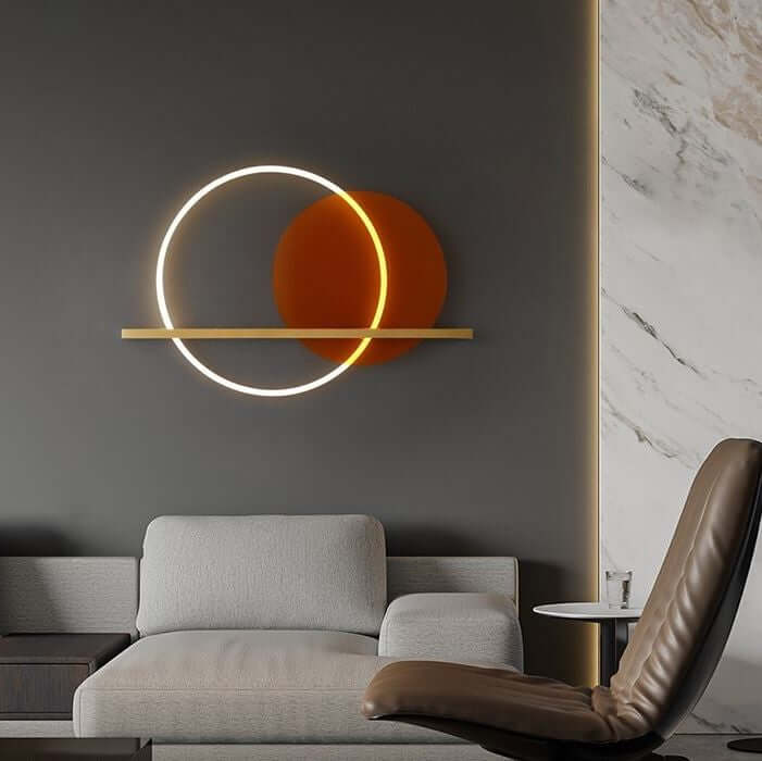 A modern wall light fixture with a brass finish and an orange acrylic circle, designed to provide ambient lighting, mounted on a wall above a grey sofa and a brown armchair.