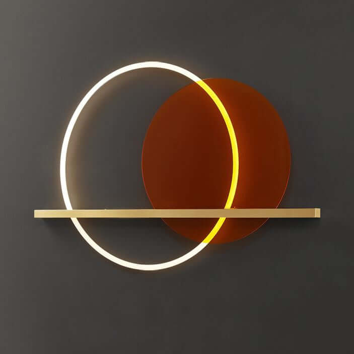 A modern wall light fixture with a brass finish and an orange acrylic circle, designed to provide ambient lighting, mounted on a wall above a grey sofa and a brown armchair.