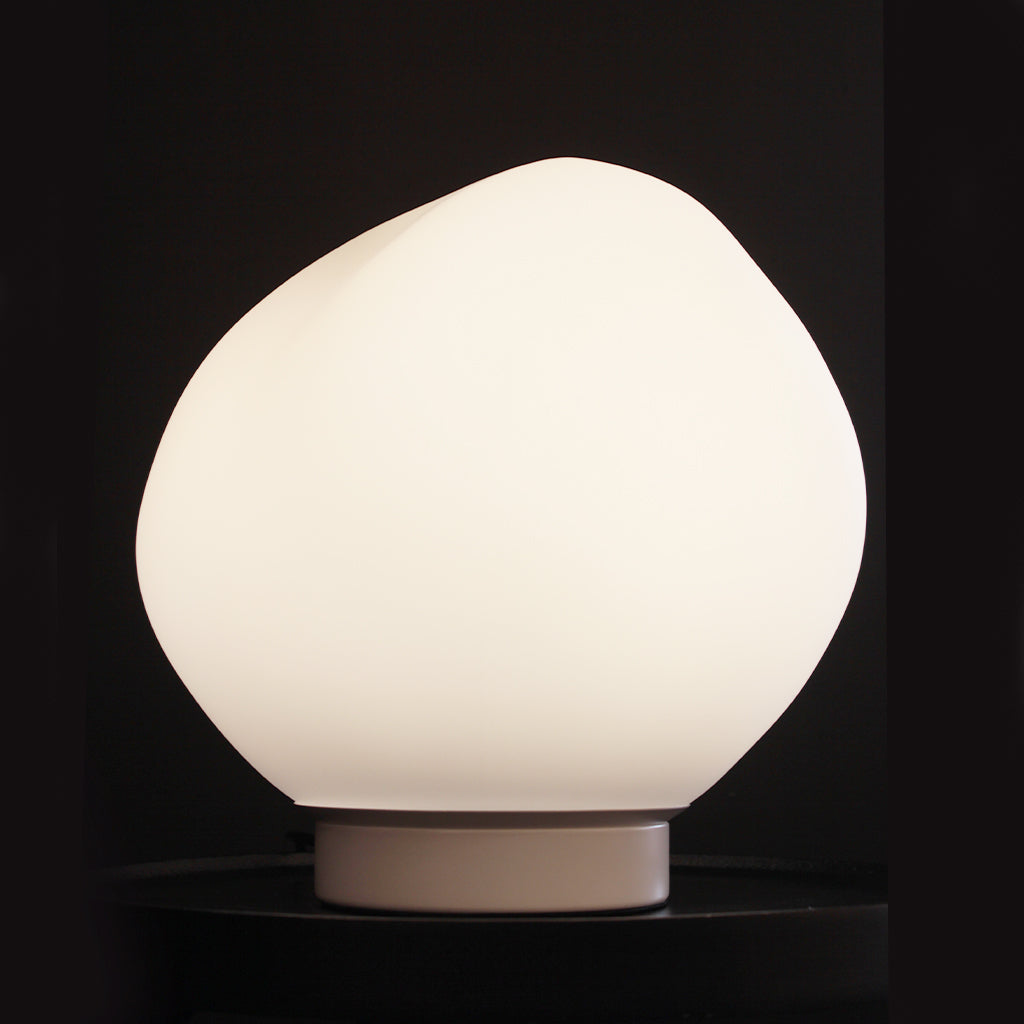 An organic-shaped matte white table lamp glowing with a warm light, resting on a matching white cylindrical base against a dark background.