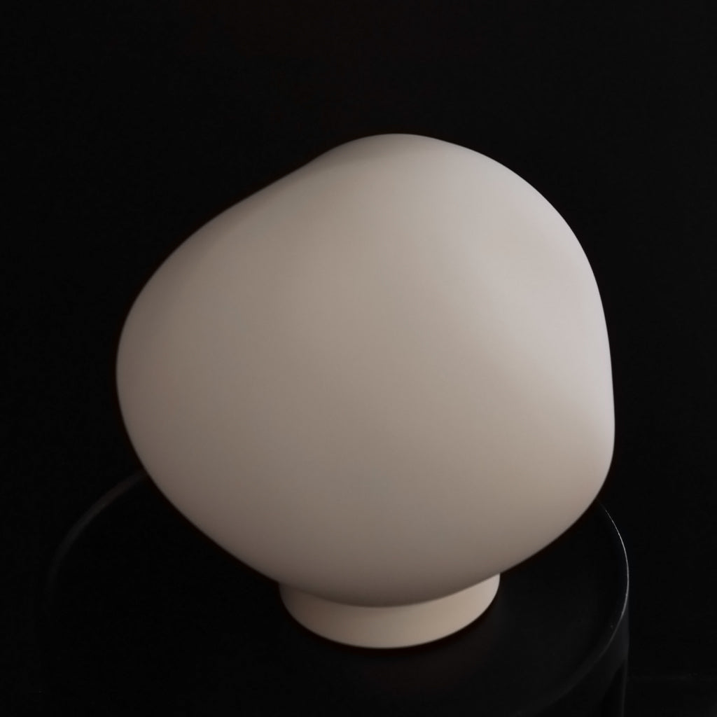 A matte off-white glass sculptural table lamp with a rounded organic shape and short cylindrical base, displayed on a black reflective surface against a black background.