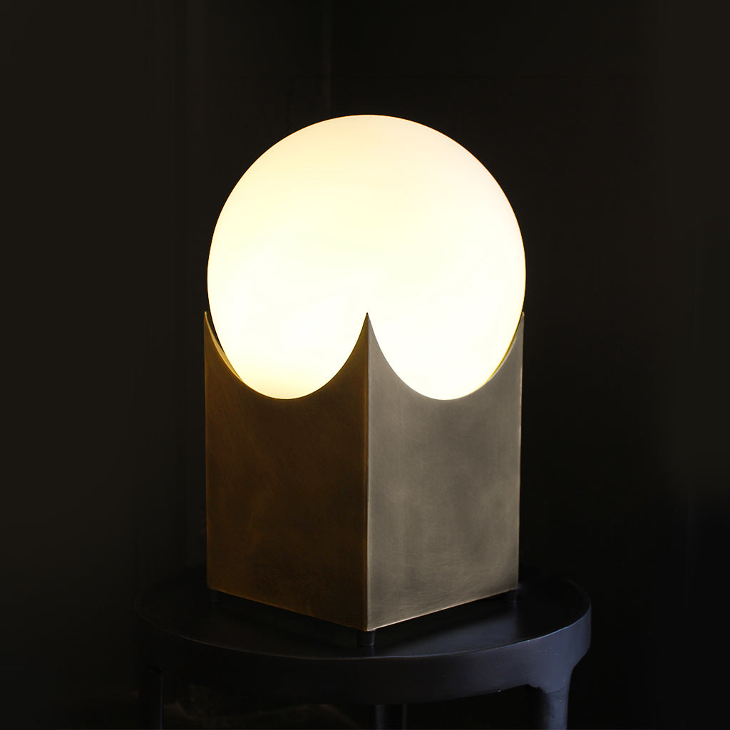 A modern table lamp with a glowing frosted oval shade and a sculptural metallic base, shown on a dark surface against a black background.