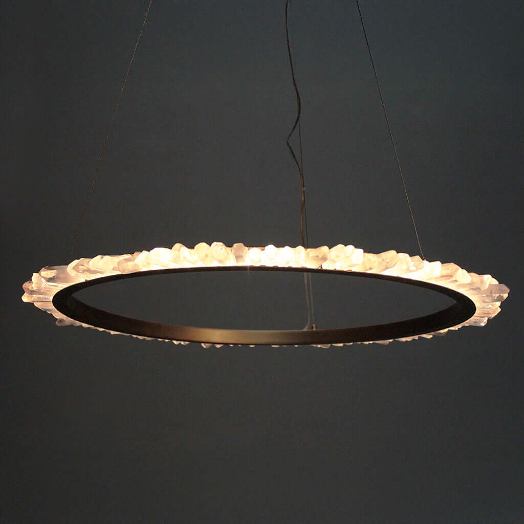 A modern circular LED pendant light with a dark Brass metal frame and a textured, natural quartz outer ring that glows warm white, suspended against a dark gray background.