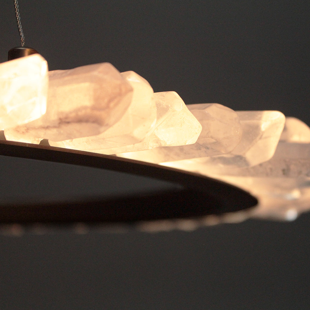 A close-up of a modern chandelier featuring a curved brass metal frame adorned with glowing, faceted clear crystal prisms that emit a warm yellow light against a dark background.
