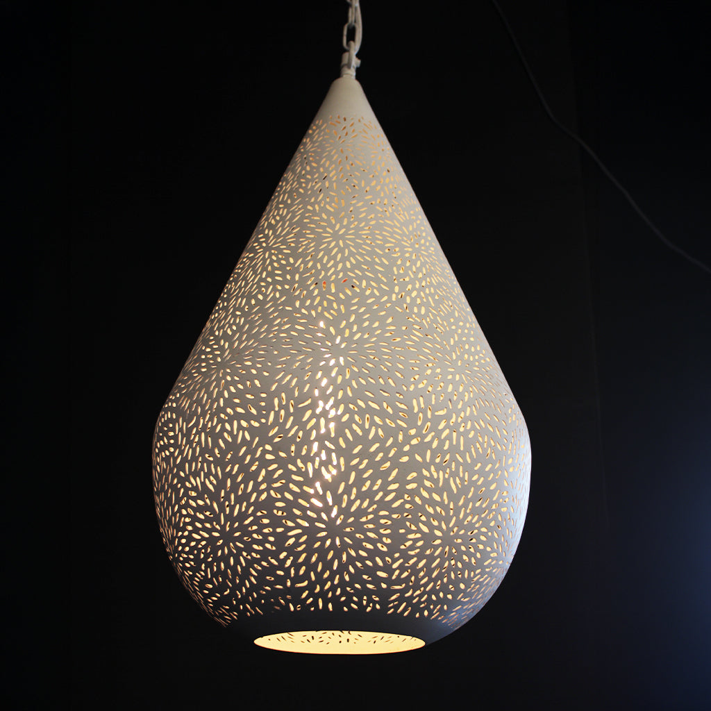 A teardrop-shaped pendant light with a white metal body and intricate laser-cut starburst patterns, glowing with a warm yellow light against a solid black background.