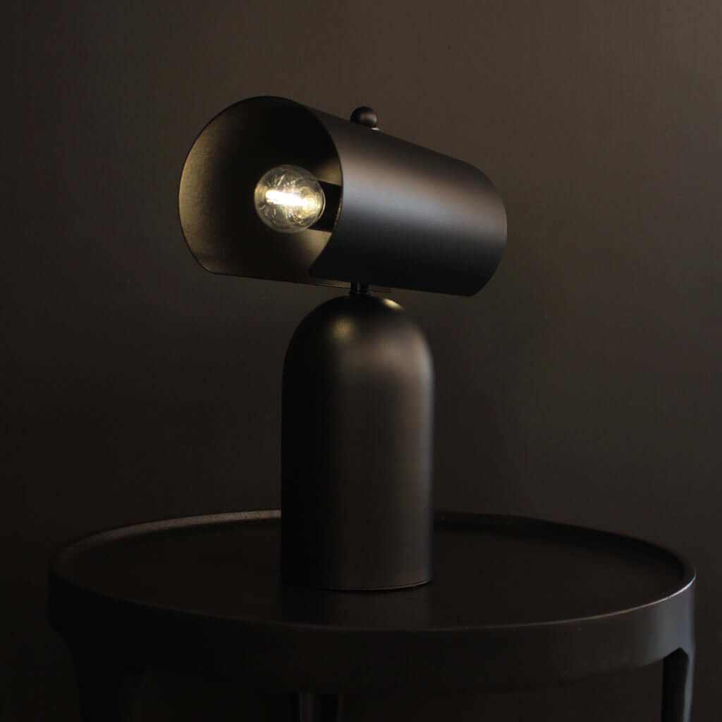 A matte black modern table lamp with a cylindrical shade and visible bulbs, shown lit on a round black side table against a dark brown background.