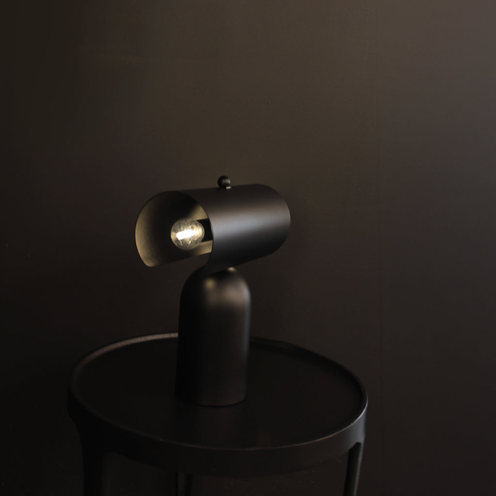 A matte black modern table lamp with a cylindrical shade and visible bulbs, shown lit on a round black side table against a dark brown background.