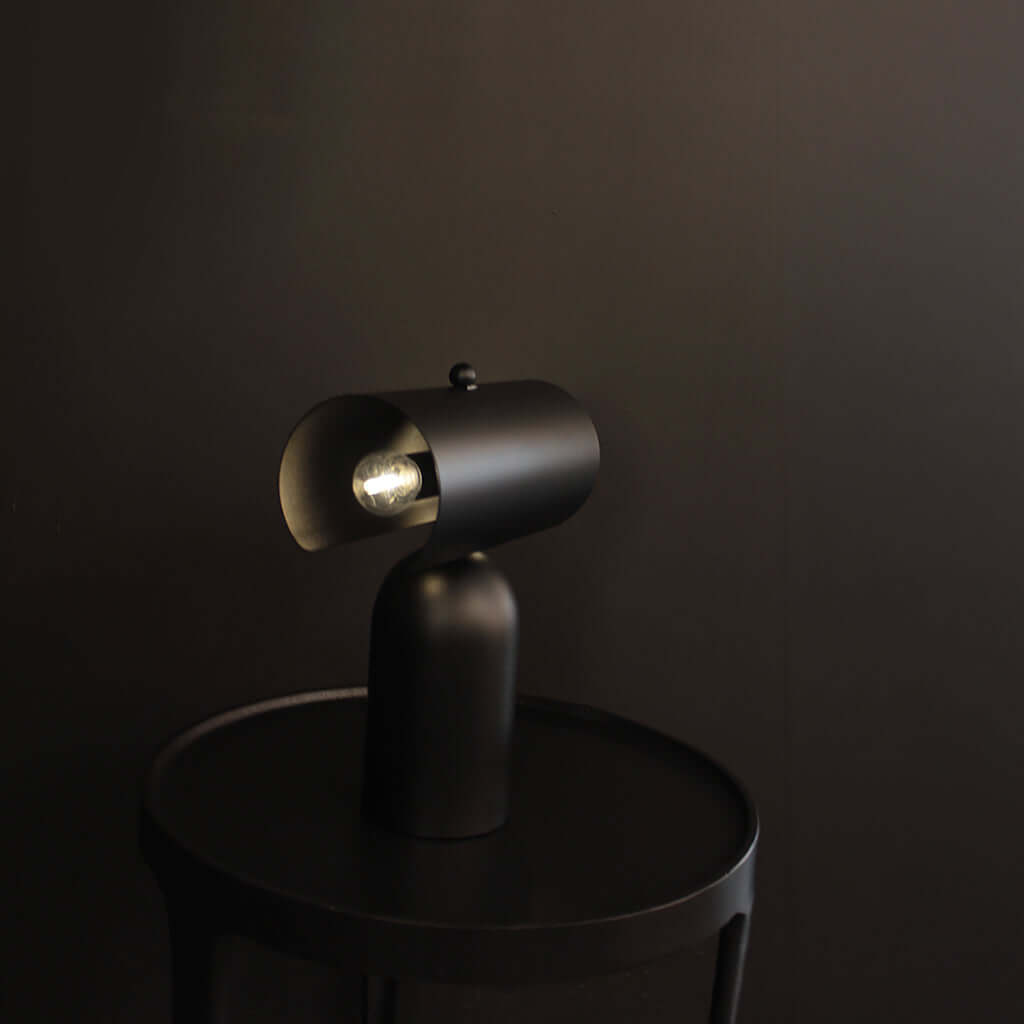 A matte black modern table lamp with a cylindrical shade and visible bulbs, shown lit on a round black side table against a dark brown background.