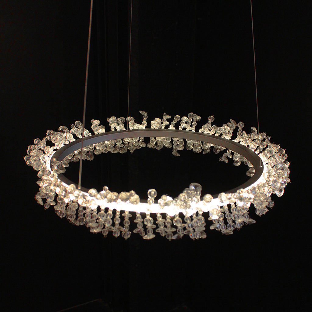 A modern circular chandelier with a silver metal ring and clear crystal beading, illuminated against a solid black background.