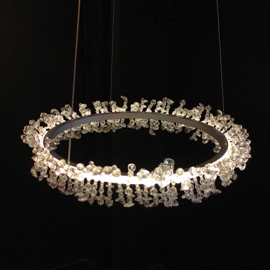 A modern circular chandelier with a silver metal ring and clear crystal beading, illuminated against a solid black background.