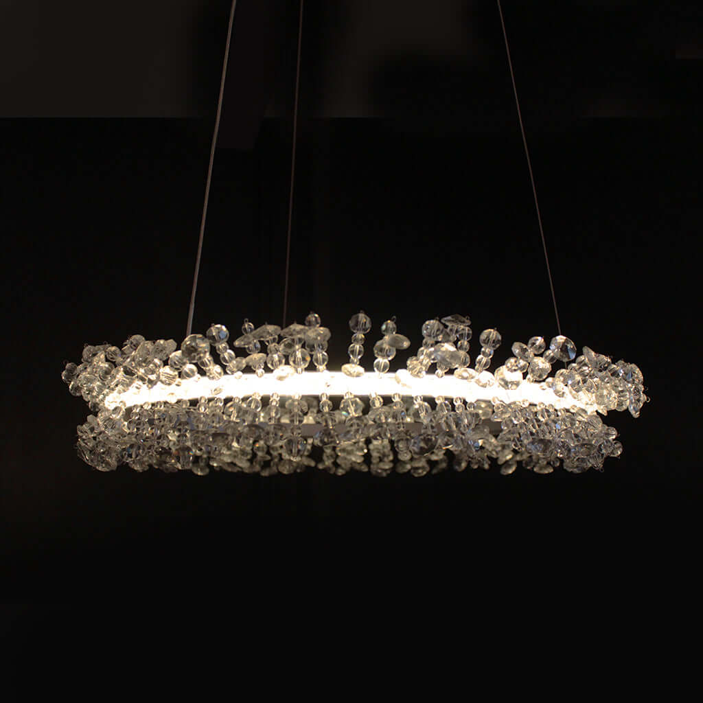 A modern horizontal crystal chandelier with a glowing white LED light ring and clear faceted crystal beads, suspended against a solid black background.