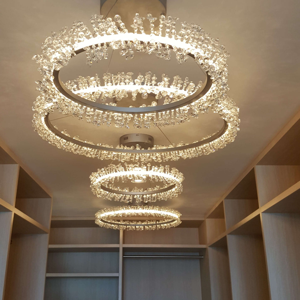 A modern multi-ring chandelier with clear crystal droplets and warm white lighting, mounted on a ceiling in an interior room with shelving.