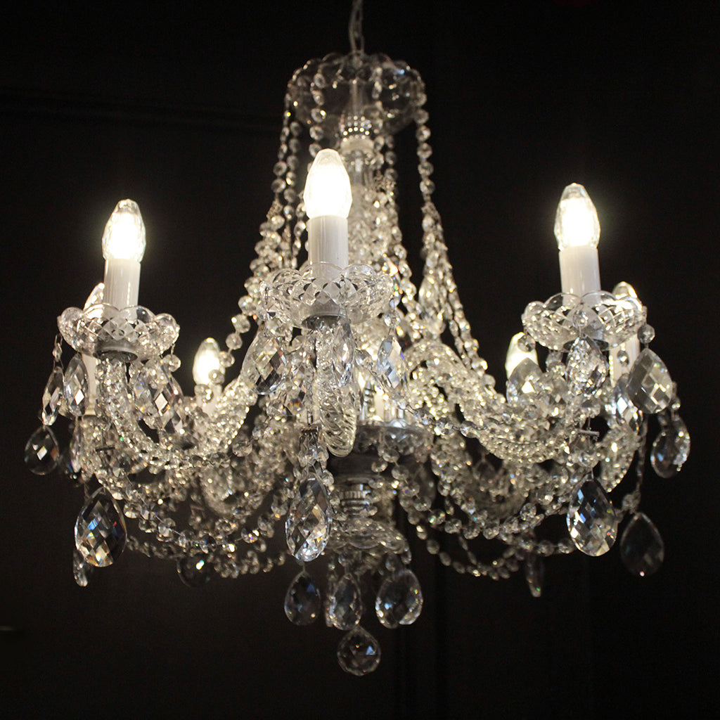 An eight-light crystal chandelier with clear faceted crystal beads, teardrop pendants, and candle-style bulbs turned on against a black background.