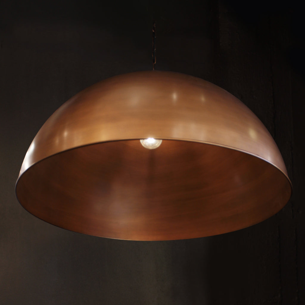 A large dome-shaped pendant light with a metallic dark brass finish, suspended against a dark background with a single bulb glowing inside.
