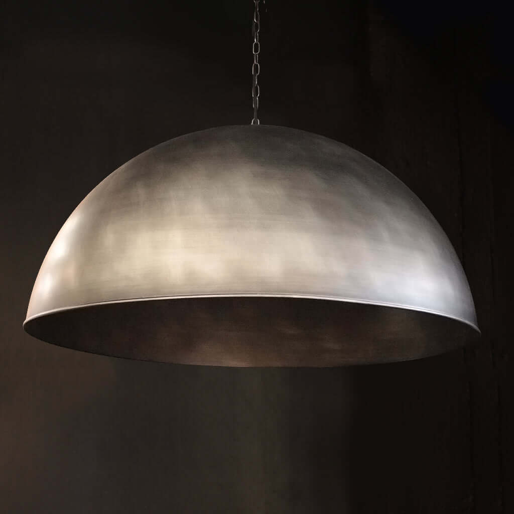 A large dome-shaped pendant light with a brushed metallic silver finish, suspended by a chain against a dark background.