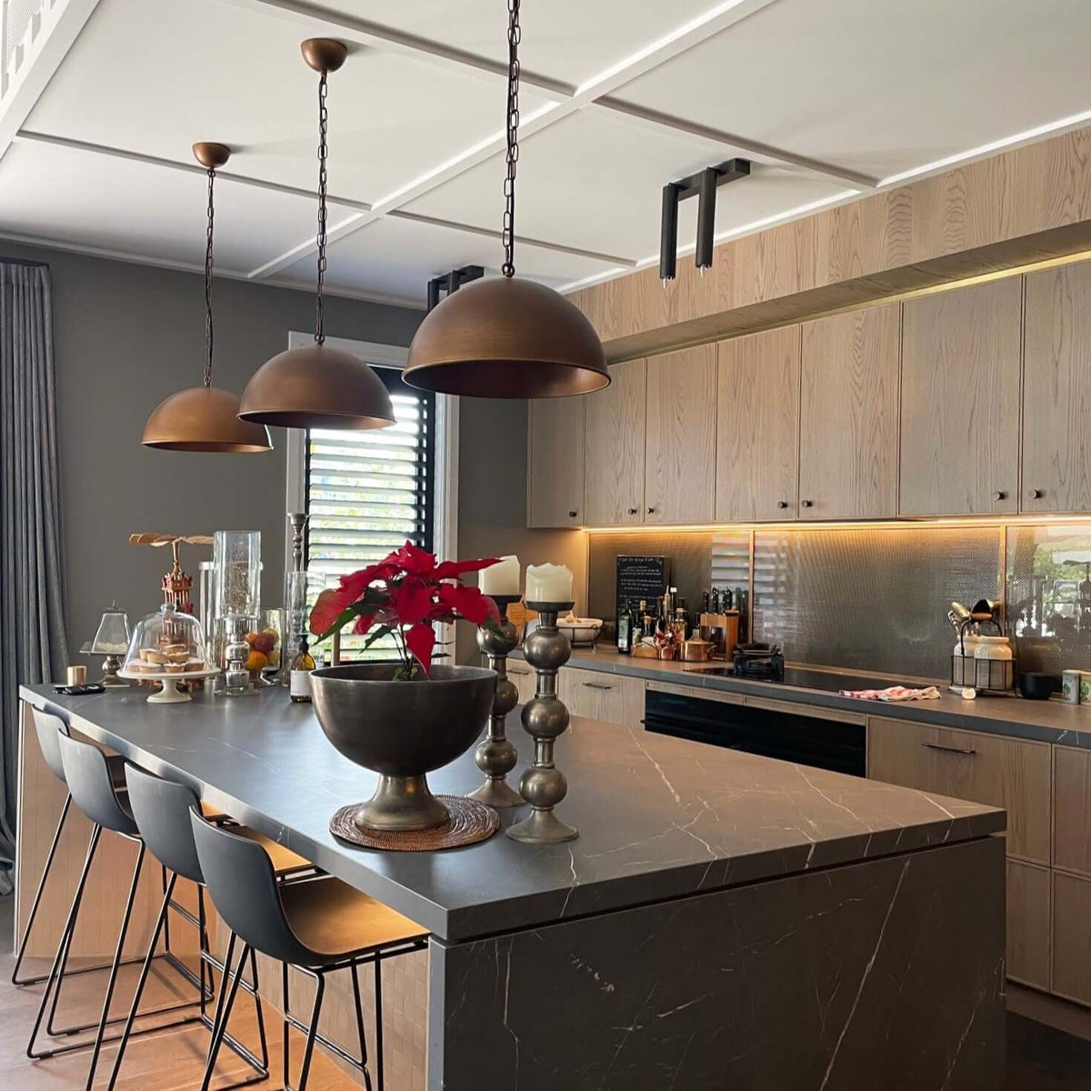 A modern kitchen interior with a large island, dark gray marble countertop, light wood cabinetry, three bronze pendant lights, and a red poinsettia arrangement on the counter.