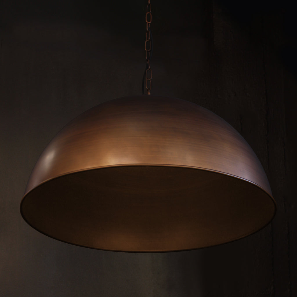 A large dome-shaped pendant light with a warm metallic brass bronze finish, suspended by a chain against a dark textured background.