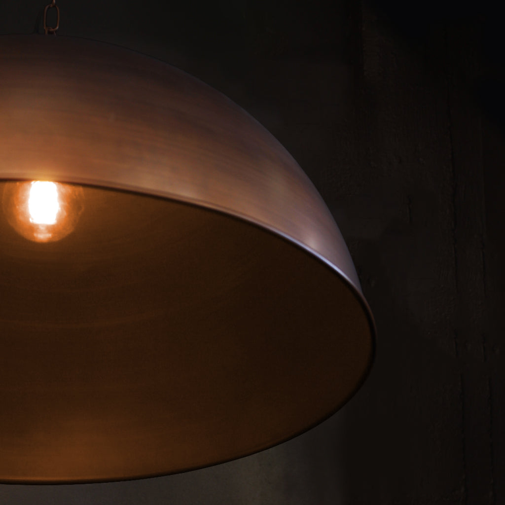 A close-up of a large dome-shaped pendant light with a warm metallic brass bronze finish, showing the glowing light bulb inside against a dark background.