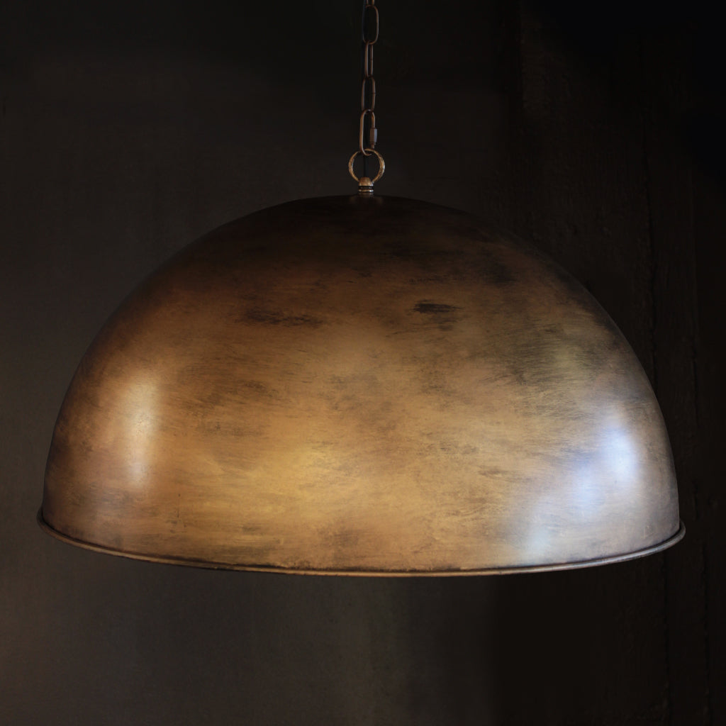 A large dome-shaped pendant light with a textured metallic brass finish, suspended by a thin chain against a dark, moody background.