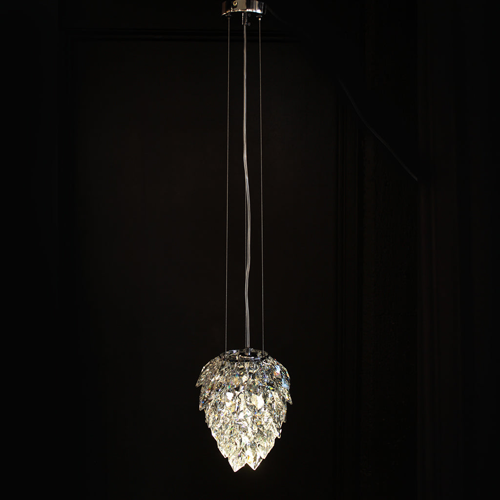 A modern pendant light fixture with a clear faceted crystal droplet-shaped shade hanging against a dark black background.