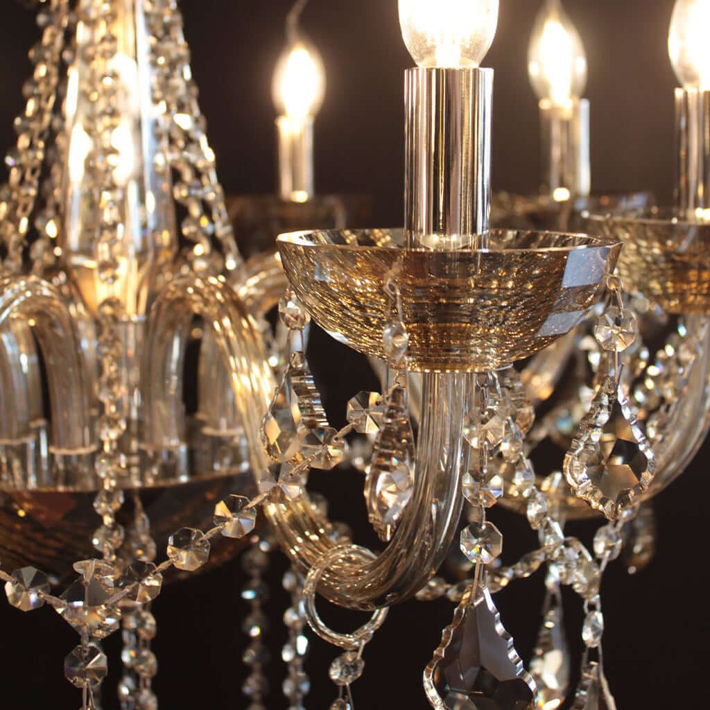 Close-up of a chandelier with amber glass crystal drops, curved amber glass arms, and gold-toned bowls, shown lit against a dark background.