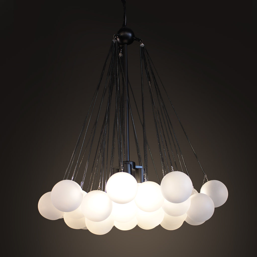 A modern multi-light pendant chandelier featuring multiple frosted white spherical glass shades suspended by thin black cords from a black central frame, shown against a dark gradient background.