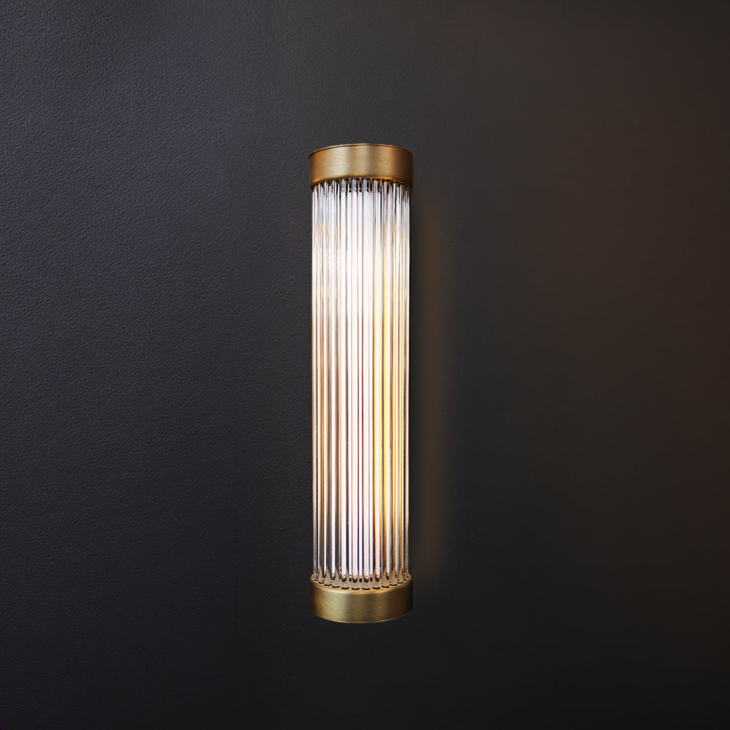 Pillar wall light with antique brass fixtures and clear glass vertical rods against dark background