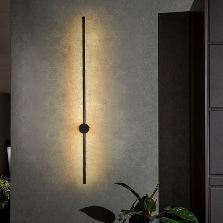 A modern minimalist wall sconce with a slim vertical black metal rod and a central circular accent, casting a warm yellow light onto a textured gray wall in an interior setting.