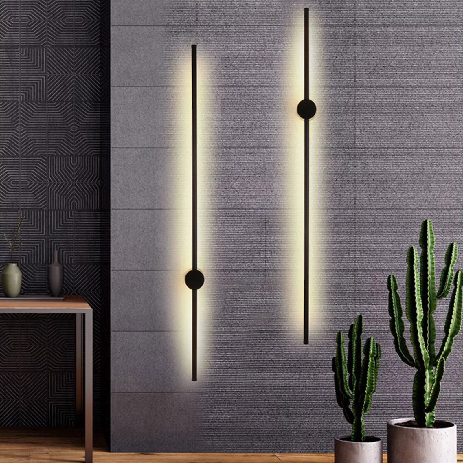 A modern minimalist wall sconce with a slim vertical black rod and a brushed gold circular accent, casting a warm yellow light on a dark gray wall in a contemporary living room setting.