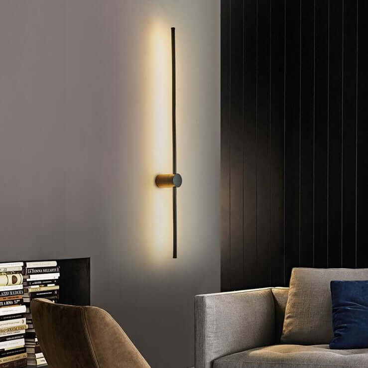 A modern minimalist wall sconce with a slim vertical black rod and a brushed gold circular accent, casting a warm yellow light on a dark gray wall in a contemporary living room setting.