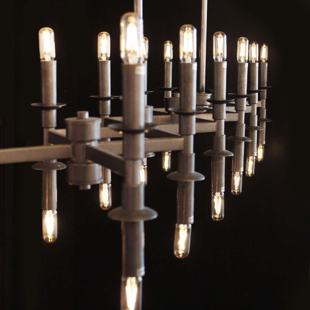 Industrial-style chandelier with a matte gray metal frame and multiple candle-shaped bulbs glowing with warm light, set against a solid black background.