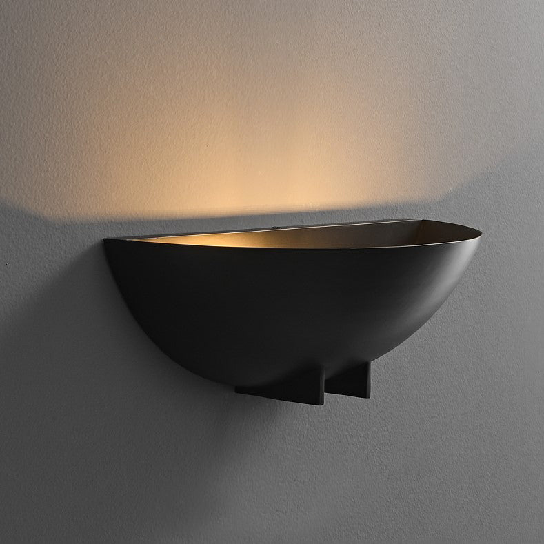 A minimalist half round wall sconce with a warm light, finished in either brushed brass or blackened iron, mounted on a wall.