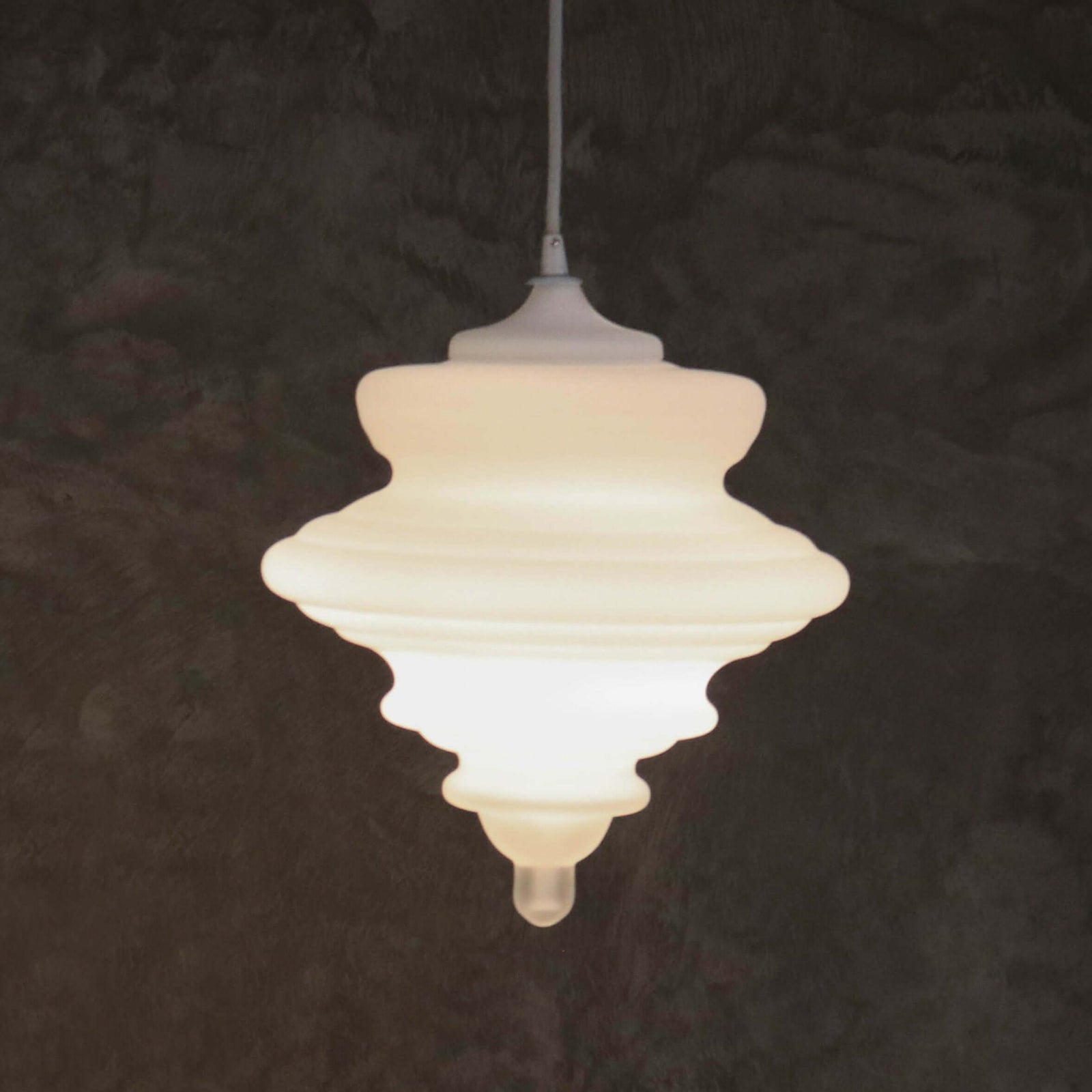 A modern, tiered pendant light with a glowing white frosted glass shade, suspended by a thin cord against a dark, textured background.