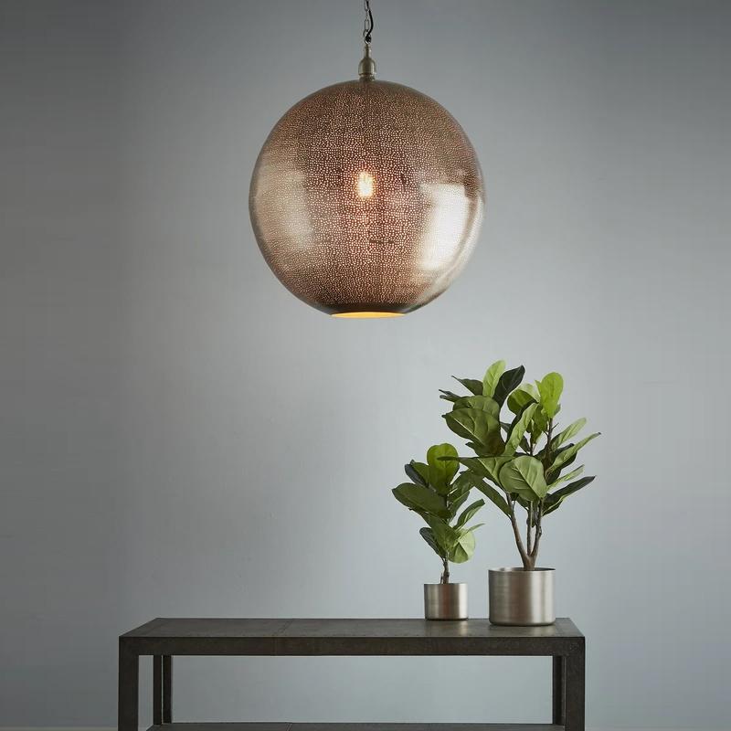 A modern pendant light with a nickel finish and small perforations in the shade, suspended over a grey table, featuring two plants and a geometric metal leg.