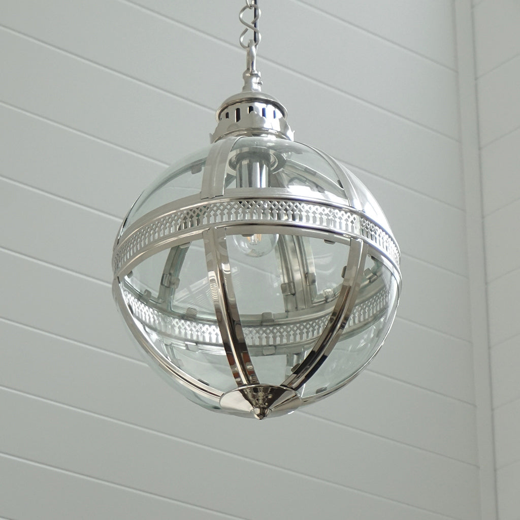A spherical pendant light fixture with a clear glass globe and polished silver metal frame, hanging against a white shiplap wall.