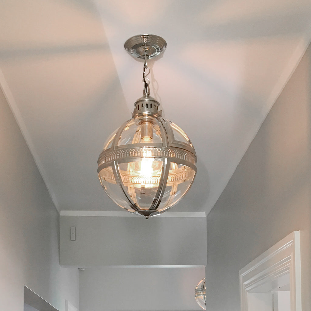 A modern spherical pendant light fixture with a polished silver metal frame and clear glass globe shade, hanging from a matching ceiling canopy in a hallway.