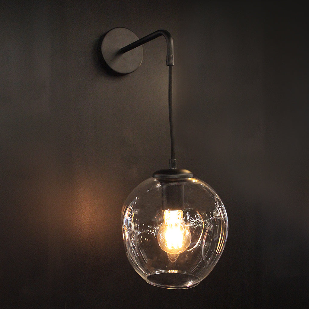 A modern wall-mounted pendant light with a Clear glass globe shade and a dark metal wall plate and cord, shown against a dark wall with the light turned on.