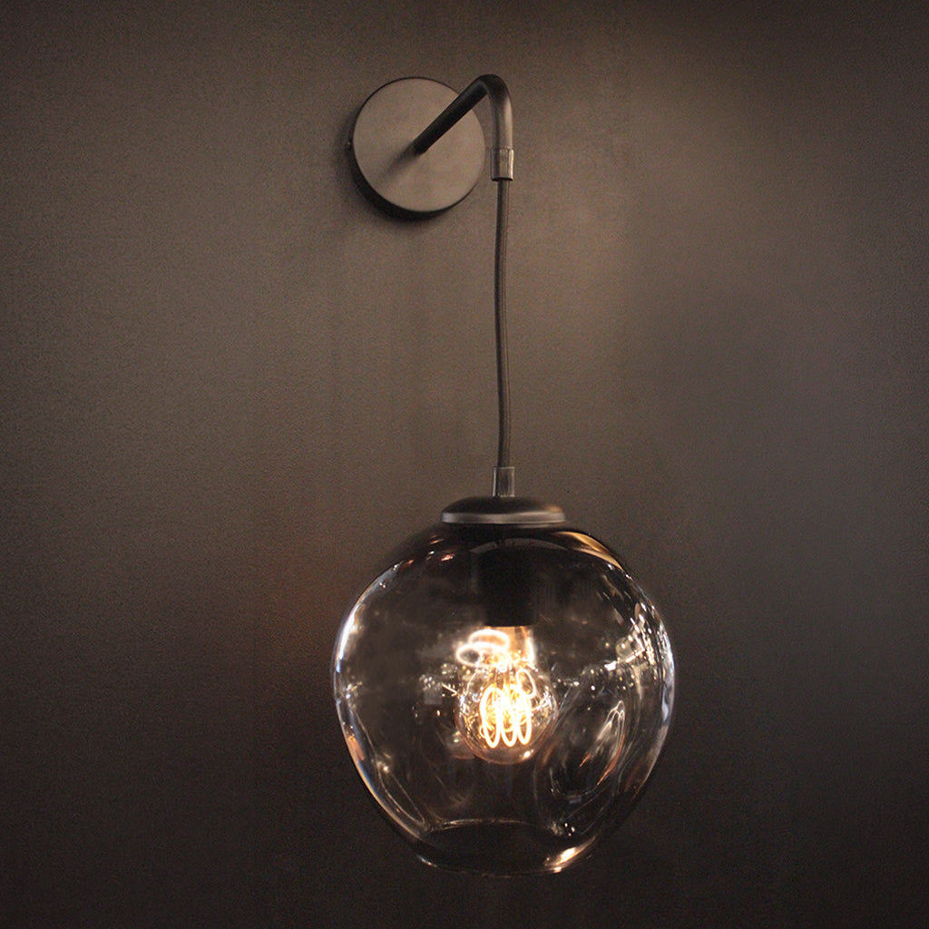 A modern wall-mounted pendant light with a Smoke Grey glass globe shade and a dark metal wall plate and cord, shown against a dark wall with the light turned on.