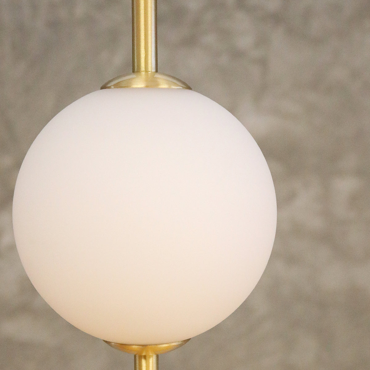 A close-up of a modern pendant light featuring a matte white spherical glass shade suspended from a polished gold-toned brass rod against a textured gray wall.
