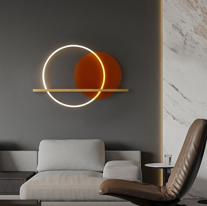 A modern wall light fixture with a brass finish and an orange acrylic circle, designed to provide ambient lighting, mounted on a wall above a grey sofa and a brown armchair.