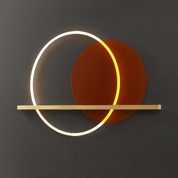 A modern wall light fixture with a brass finish and an orange acrylic circle, designed to provide ambient lighting, mounted on a wall above a grey sofa and a brown armchair.