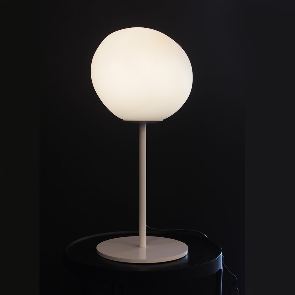 A modern spherical table lamp with a glowing white globe shade and white base, placed on a dark wooden side table next to a striped armchair in a dimly lit room.