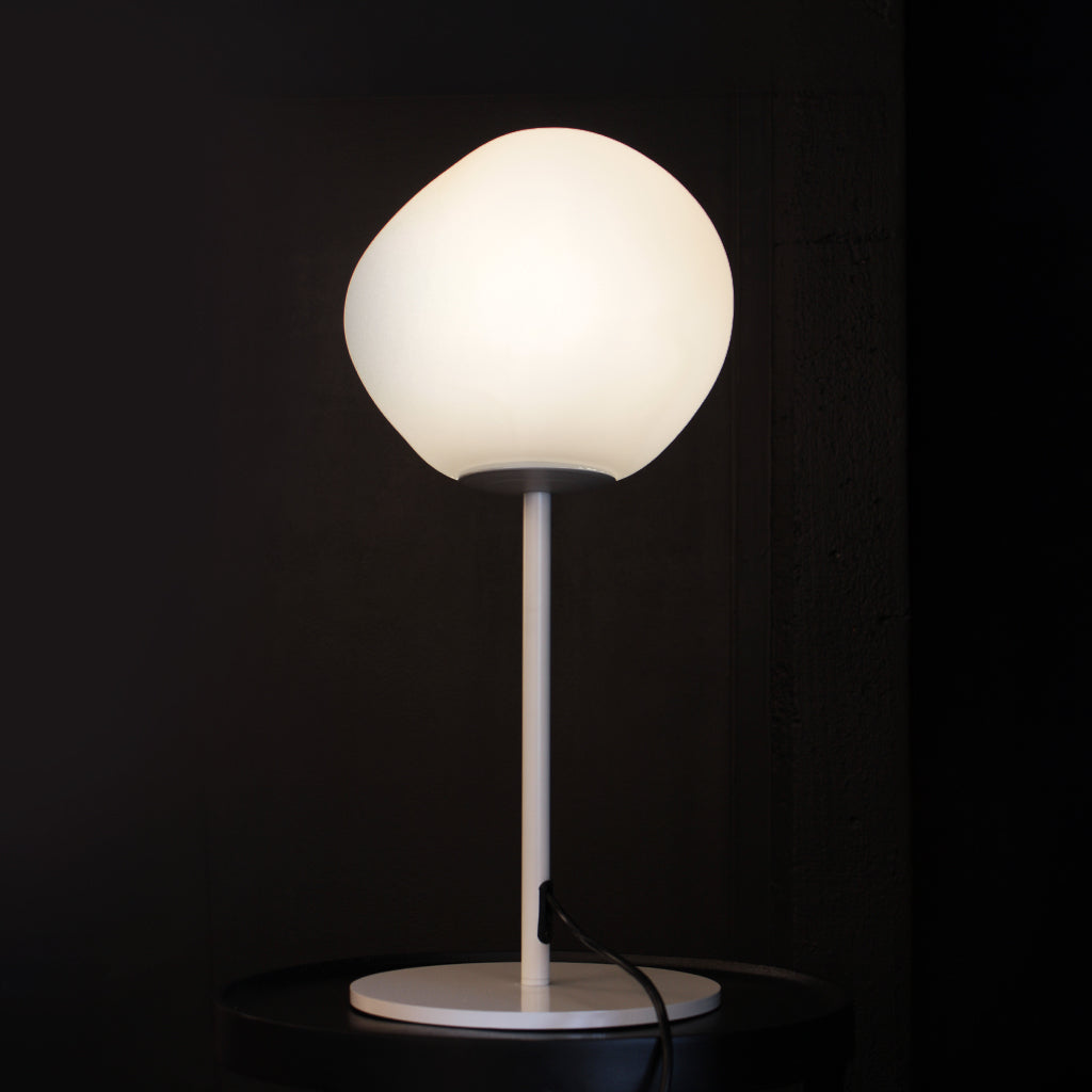 A modern table lamp with a glowing white organic-shaped globe shade, a slim white stem, and a round white base, set against a dark background.