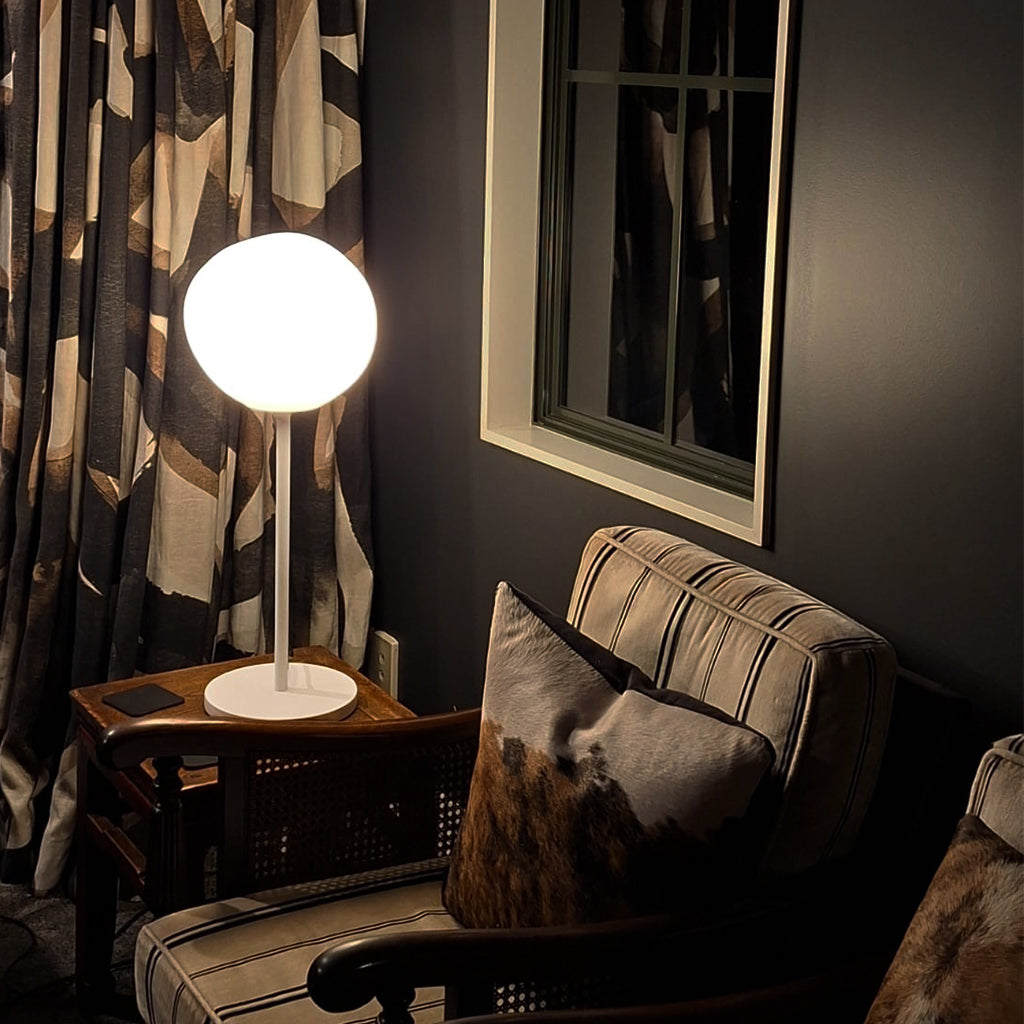 A modern spherical table lamp with a glowing white globe shade and white base, placed on a dark wooden side table next to a striped armchair in a dimly lit room.