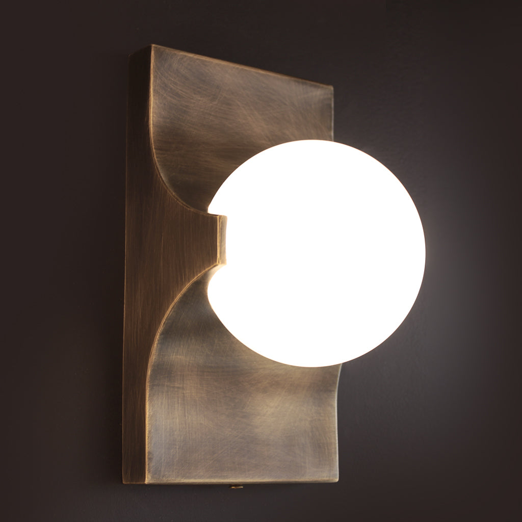 A modern wall-mounted light fixture with a brushed brass or bronze-toned metal frame and a glowing white spherical shade, shown against a dark wall.