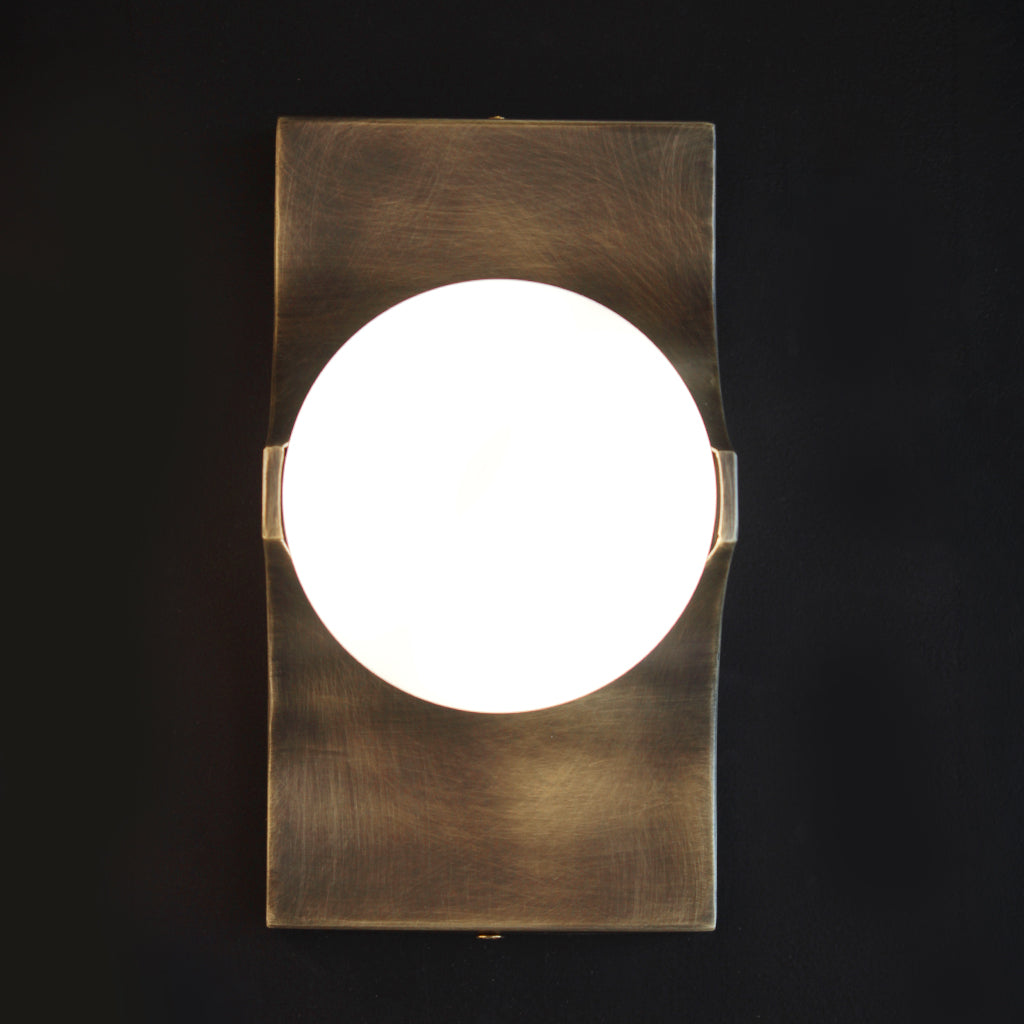 A modern wall-mounted light fixture with a brushed brass or bronze-toned metal frame and a glowing white spherical shade, shown against a dark wall.
