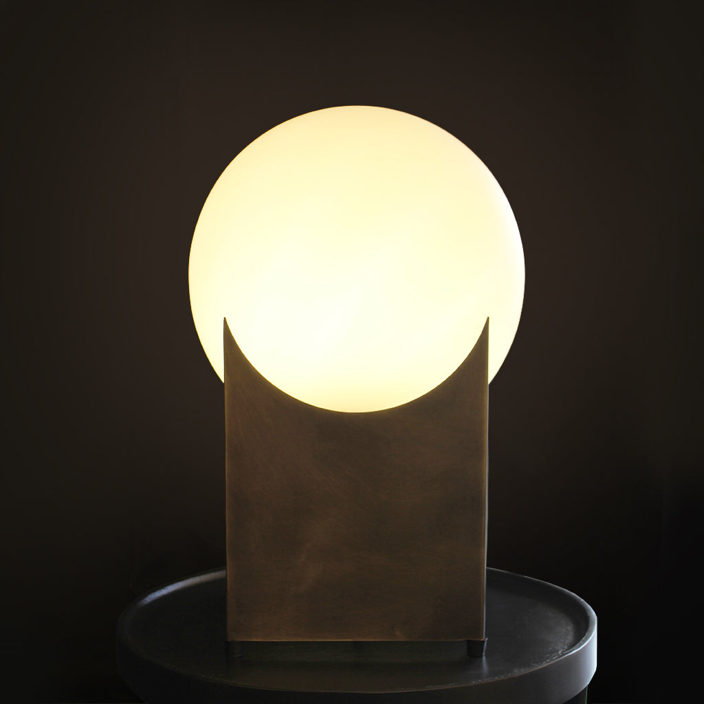 A modern table lamp with a glowing round frosted shade and a dark textured geometric base, placed on a wooden table in a softly lit interior room.